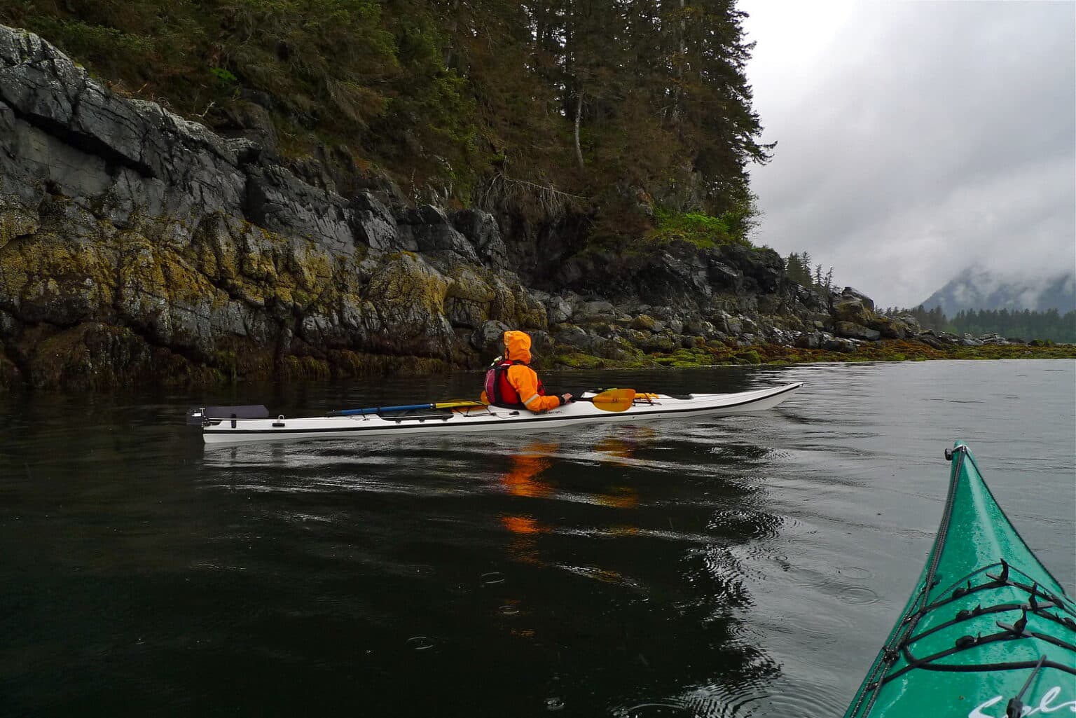 Kayaker in vibrant orange gear paddling near a rocky shoreline with misty mountains in the background