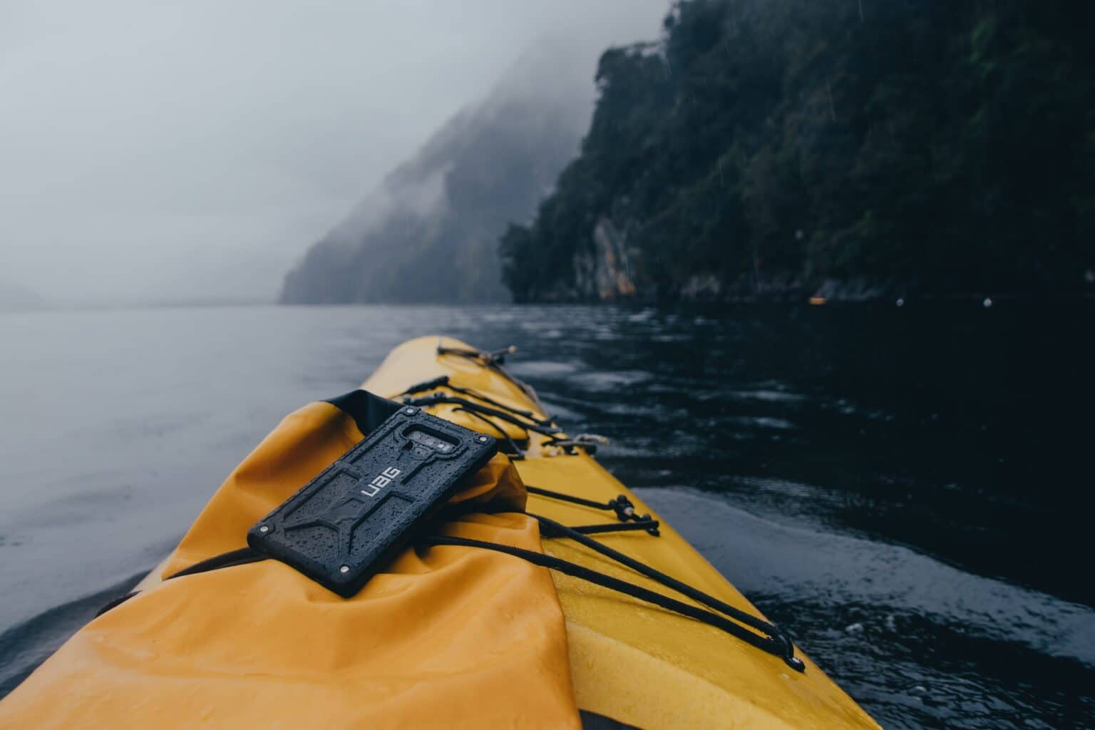 Yellow kayak on calm water with waterproof gadget against misty mountain backdrop